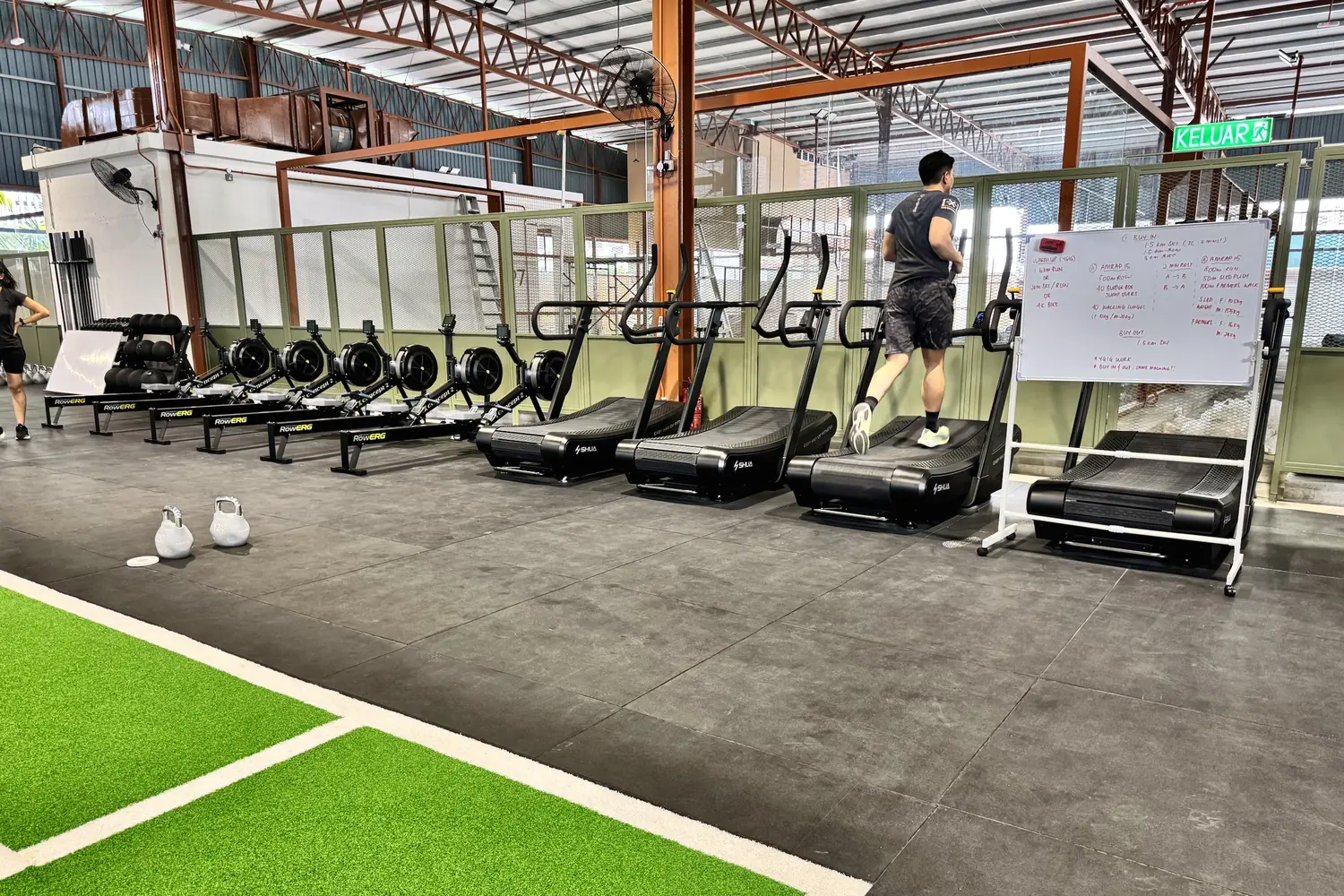 A row of rowers and curved treadmills, one member running, the day's workout on a whiteboard