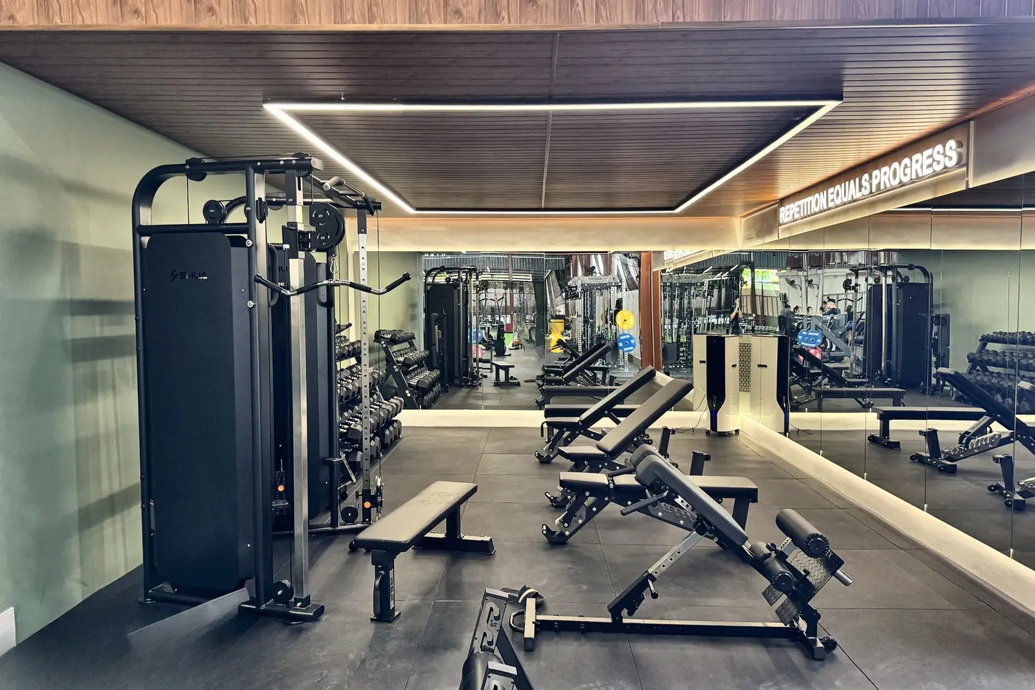 The strength room: a cable station, benches and dumbbells under a lit timber ceiling, mirrors along one wall